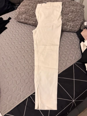 Vita raka chinos med dragkedja - Säljer ett par stilrena vita chinos med rak passform och dragkedja vid benslut. Byxorna har klassiska fickor och bälteshällor i midjan. Perfekta för en clean och fräsch look. Materialet känns mjukt och lätt, troligen bomull eller bomullsblandning.