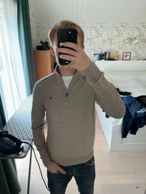 Beige halvziptröja från Morris - Snygg beige halvziptröja från Morris med hög krage och diskret broderad logga på bröstet. Tröjan är tillverkad i mjukt material och har ribbade muddar vid ärmslut och nederkant. Perfekt för lager på lager och stilrena outfits.