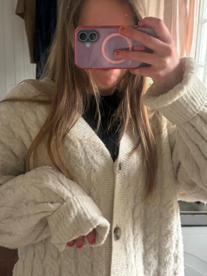 Vit kabelstickad kofta med knappar - Mysig vit kabelstickad kofta med stora knappar framtill och ribbade muddar. Koftan har en oversized passform och är perfekt för lager på lager. Tjockt material som håller dig varm under kyliga dagar.