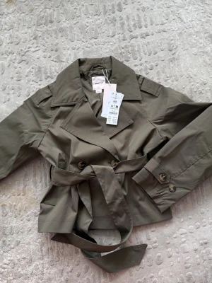 Kort khakigrön trenchcoat med bälte - Stilren kort trenchcoat från Gina Tricot i khakigrönt bomullstyg. Jackan har bred krage, dubbelknäppning framtill, axelklaffar och ett matchande knytbälte i midjan. Ärmarna har dekorativa knappar vid mudden och jackan har en klassisk stormflik bak.