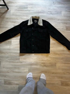 Svart jeansjacka med teddykrage - Snygg svart jeansjacka från Divided med vit teddykrage. Klassisk modell med två bröstfickor med lock och knappar framtill. Jackan har raka ärmar och en avslappnad passform, perfekt för en cool och trendig look.
