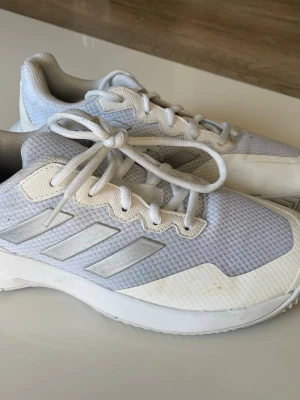 Vita padelskor från Adidas - Säljer ett par vita padelskor från Adidas med klassiska tre ränder på sidan. Skorna har mesh-ovandel för bra ventilation och vita snören. Snygg och sportig design som passar perfekt till träning eller streetwear.