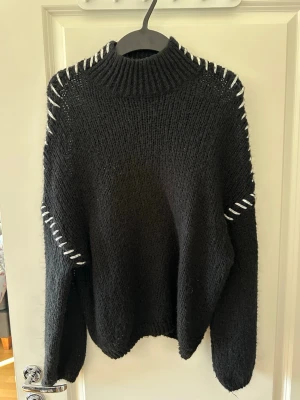 Svart stickad tröja med vita detaljer - Säljer en svart stickad tröja med höghals och vita dekorativa sömmar längs axlar och ärmar. Tröjan har en avslappnad passform och långa ärmar. Perfekt för dig som gillar en enkel men ändå unik look.