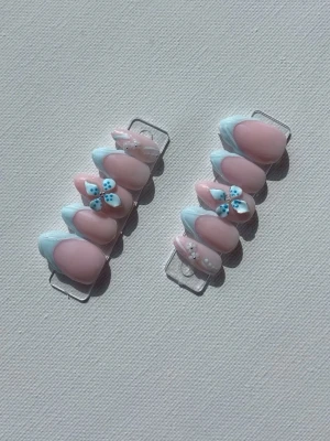 Press-on naglar med blå blommor - Snygga press-on naglar i mandelform med ljusrosa bas och ljusblå fransk tipp. Några naglar har 3D-dekorationer i form av blå blommor och små pärlor för extra detalj. Perfekt för dig som vill ha en söt och trendig look.