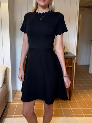 Svart kortklänning med vågig kant - Supersnygg svart kortklänning med rund hals och vågig kant vid halsringning och ärmslut. Den är från By Clara 🩷Storlek S/M. Använd en gång