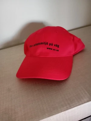Neonrosa keps med tryck - Snygg neonrosa keps med svart text framtill där det står 'Bra arbetsmiljö på väg' och 'www.av.se'. Klassisk böjd skärm och justerbar passform. Tillverkad i lätt och slitstarkt material, perfekt för dig som vill sticka ut.