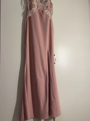 Rosa långklänning med spetsdetaljer - Elegant rosa långklänning med v-ringning och vita spetsbroderier över byst och midja. Klänningen har ärmlös design och figurnära passform upptill, vilket ger en romantisk och feminin look. Perfekt för speciella tillfällen.