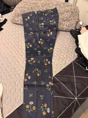 Mörkblå kostymbyxor med blommor - Snygga mörkblå kostymbyxor med färgglatt blommönster i gult, lila och grönt. Byxorna har raka ben, bälteshällor och fickor bak. Materialet ser ut att vara glansigt och tunt, vilket ger en elegant känsla. Perfekta för dig som vill sticka ut med din stil.