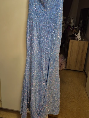 Blå glittrig långklänning med slits - Säljer en blå långklänning täckt av glittriga paljetter som ger ett skimrande intryck. Klänningen har en elegant slits framtill och figurnära passform. Perfekt för dig som vill sticka ut på bal eller fest.