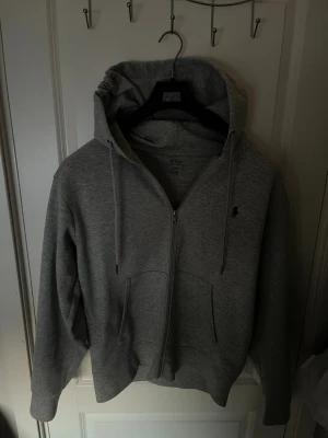 Grå hoodie från Polo Ralph Lauren - Klassisk grå hoodie från Polo Ralph Lauren med dragkedja framtill, två fickor och justerbar huva med snörning. Liten broderad logga på bröstet. Perfekt för en chill och stilren look. Kan gå ned lite vid snabb affär 