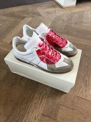 Japan gats  - Unika sneakers från Maison Margiela i vitt och grått läder med en stor röd cirkel på sidan och matchande röda skosnören. Klassisk low-top siluett och gummisula. Perfekta för dig som vill sticka ut med en cool och modern look.