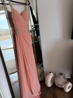 Lång rosa balklänning med pärldetaljer - Jovani  långklänning i ljusrosa med draperad överdel och transparent mesh upptill. Klänningen har glittriga pärldetaljer längs axelband och sidor, samt en figurnära passform. Perfekt för bal eller festliga tillfällen.