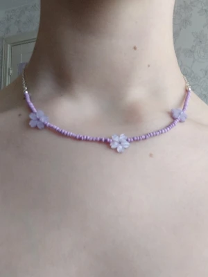 Lila pärlhalsband med blommor - Ett sött halsband med små lila pärlor och tre blommor i plast. Halsbandet har en tunn kedja i silverfärg vid låset och resten består av lila pärlor. Perfekt för att ge en lekfull touch till din outfit.