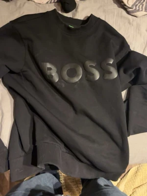 Svart hoodie från BOSS - Snygg svart hoodie från BOSS med stor logga framtill. Klassisk modell med rund hals och långa ärmar. Perfekt för dig som gillar stilrena och trendiga plagg med tydlig branding.