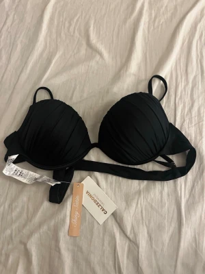 Svart bikiniöverdel från Calzedonia - Stilren svart bikiniöverdel från Calzedonia i glansigt satinmaterial. Bikinin har vadderade kupor och smala axelband samt knyt i ryggen för justerbar passform. Perfekt för en trendig strandlook i sommar!