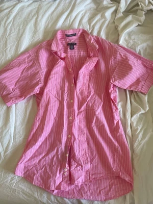 Rosa randig skjorta från GANT storlek M - Säljer en rosa skjorta från GANT med vita tunna ränder. Skjortan har korta ärmar, klassisk krage och knäppning framtill. Tillverkad i 100% bomull vilket gör den skön och luftig. Perfekt för dig som gillar en avslappnad men ändå stilren look. Storlek M