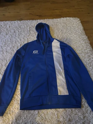 Blå hoodie från Pro Touch med vit detalj - Snygg blå hoodie från Pro Touch med dragkedja och huva. Framsidan har en vit vertikal rand och logga på bröstet. Tillverkad i ett mjukt, sportigt material som passar perfekt till träning eller chill. Två fickor framtill och ribbade muddar.