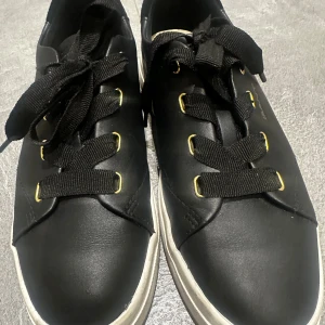 Svarta sneakers från Gant - Snygga svarta sneakers från Gant med gulddetaljer och vita sulor. Skorna har klassisk låg design, svarta breda skosnören och är tillverkade i slätt läder. 