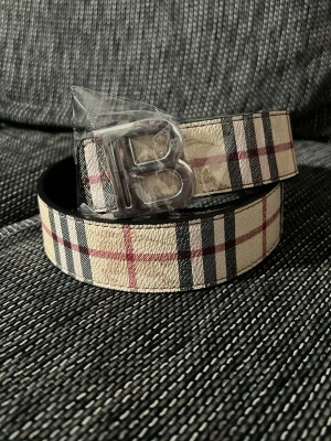  Burberry  bälte  - Snyggt skärp med klassiskt rutigt mönster i beige, svart och rött. Skärpet har ett stort silvrigt B-format spänne som ger en cool detalj. Perfekt accessoar för att lyfta din outfit.