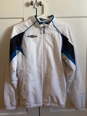 Vit träningsjacka från Umbro - Snygg vit träningsjacka från Umbro. Lite tunnare material men ändå varm, nät inuti, och dragkedja framtill, hög krage och elastiska muddar vid ärmslut och nederkant. Perfekt för träning eller sportiga outfits.     •Märke umbro                                                  •Köpt för 300/400kr                                      •Mitt pris 200kr