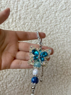 Blå fjärilsnyckelring med pärlor - Säljer en glittrig nyckelring med en stor blå fjäril dekorerad med strass och pärlor i blått, vitt och silver. Metallfäste upptill för enkel fastsättning på väska eller nycklar. Perfekt accessoar för att ge din look extra bling och färg. 💛liten extra gåva vid varje köp! Skickar allt omsorgsfullt i bubbelplast & kartong