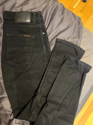 Svarta jeans från Nudie Jeans - Säljer ett par svarta jeans från Nudie Jeans med klassisk femficksdesign och diskret logga på bakfickan. Jeansen har smal passform och är tillverkade i mjukt denimtyg. Perfekta för en stilren och avslappnad look.