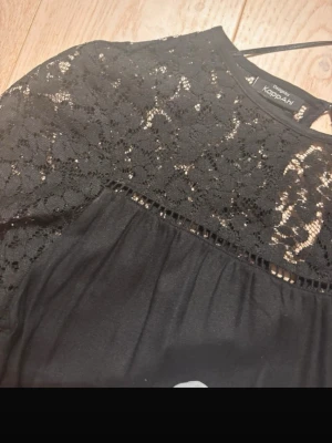 Svart spetsblus från KappAhl - Snygg svart blus från KappAhl med transparent spets upptill och över axlarna. Blusen har långa ärmar och ett lätt rynkat fall. Perfekt för dig som gillar en elegant och trendig look. Materialet är en mix av spets och mjukt tyg.