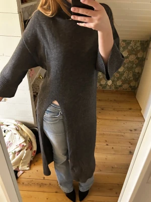 Mörkgrå stickad långtröja/klänning med slits - Säljer en mörkgrå stickad långtröja/klänning med hög krage och breda, trekvartsärmar. Tröjan har en djup slits på sidan som ger en cool och avslappnad look. Perfekt att bära över jeans eller leggings. Materialet känns mjukt och mysigt. 50% wool.