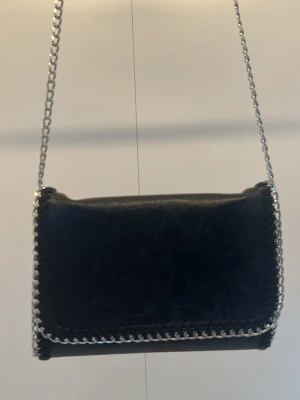 Svart axelväska med kedjerem - Snygg svart axelväska i fejkläder med silverfärgad kedjerem och dekorativ kedja runt kanten. Väskan har en stilren, rektangulär form och coolt, lite skrynkligt material. Perfekt för dig som gillar en edgy och trendig look.💞