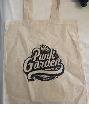 Beige tygkasse Punk Garden - Snygg beige shoppingväska i tyg från Punk Garden med svart tryck och cool logga på framsidan. Perfekt att bära till skolan, gymmet eller när du handlar. Väskan har två långa handtag och är tillverkad i slitstark bomull.