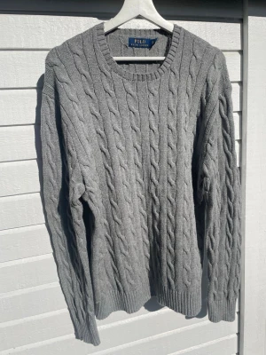 Grå kabelstickad tröja Polo Ralph Lauren - Snygg grå kabelstickad tröja från Polo Ralph Lauren i 100% bomull. Tröjan har rund halsringning, långa ärmar och ribbade muddar vid ärmslut och nederkant. Perfekt för lager på lager och en klassisk stil.
