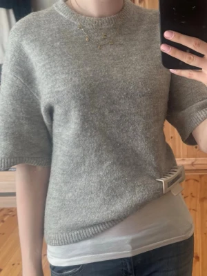 Grå stickad tröja Gina Tricot - Mysig grå stickad tröja från Gina Tricot med korta ärmar och rund halsringning. Tröjan har en enkel, rak passform och ribbade kanter vid hals, ärmslut och nederkant. Perfekt att styla med jeans eller kjol för en avslappnad look.
