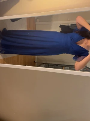 Lång blå balklänning med knyt i midjan - Elegant långklänning i djupblå färg med v-ringning och korta, fladdriga ärmar. Klänningen har ett fint knytband i midjan som framhäver siluetten och ett lätt, flytande tyg som ger en snygg drapering. Perfekt för speciella tillfällen.