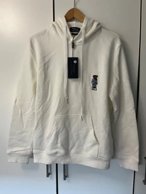 Vit hoodie från Polo Ralph Lauren - Vit hoodie från Polo Ralph Lauren med broderad ikonisk björn på bröstet. Klassisk känguruficka framtill, ribbade muddar och dragsko i huvan. Tillverkad i mjuk bomull för en skön och avslappnad look.