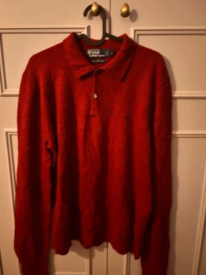 Röd ulltröja Polo Ralph Lauren - Röd långärmad tröja från Polo Ralph Lauren i 100% lammull. Tröjan har pikékrage med två knappar och en liten broderad logga på bröstet. Perfekt för dig som gillar klassisk och stilren design med en exklusiv touch.