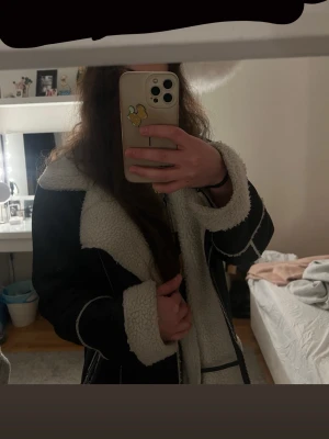 Svart pälsjacka med vit fuskpäls - Säljer en svart pälsjacka med vit fuskpäls på krage, ärmslut och insida. Jackan har en oversized passform och markerade sömmar för en trendig look. Perfekt för kalla dagar och ger en cool vibe till din outfit.