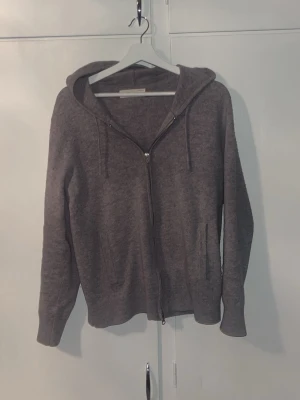 Sayless stickad tröja - Mysig mörkgrå hoodie med dragkedja framtill och två fickor. Tillverkad i mjukt material och har en klassisk huva med dragsko. Perfekt för en avslappnad stil och enkel att matcha med andra plagg.