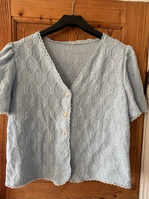 Ljusblå spetsblus med knappar - Supersöt ljusblå blus med korta puffärmar och v-ringning. Blusen har dekorativa spetsdetaljer i ett rutmönster och tre vita knappar framtill. Avslut med vågig spetskant. Perfekt till jeans eller kjol för en romantisk look.