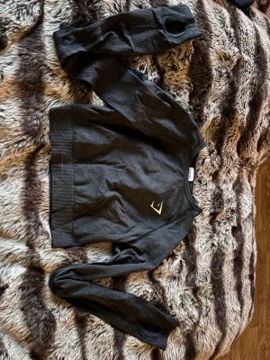 Svart croppad gymshark träningströja - Svart långärmad träningströja från Nike med diskret struktur i materialet och tumhål i ärmsluten. Tröjan har en liten gul Nike-logga på bröstet och ribbade muddar. Perfekt för löpning eller gym.