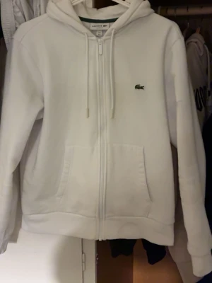 Vit zip-hoodie från Lacoste - Säljer en clean vit hoodie från Lacoste med dragkedja, huva med dragsko och klassisk krokodil-logga på bröstet. Hoodien har två stora fickor fram och är i mjukt material, perfekt för chill dagar. Classic fit-modell.