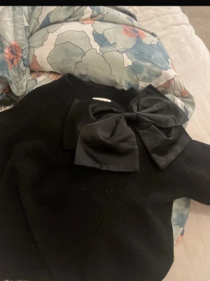 Svart stickad tröja med stor rosett - Snygg svart stickad tröja med en stor dekorativ rosett på axeln. Tröjan har rund halsringning och långa ärmar. Perfekt för dig som vill ha något extra till din outfit. (Pris kan diskuteras)