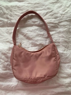 Rosa axelremsväska i nylon - Söt rosa handväska i nylon med enkel, rundad form och dragkedja upptill. Väskan har en bred axelrem och minimalistisk design som passar perfekt till en trendig vardagslook. Ingen synlig logga eller märke.