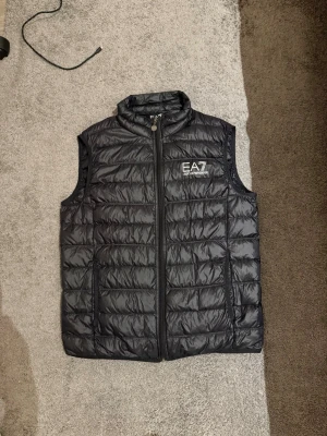Svart dunväst från EA7 Emporio Armani - Snygg svart dunväst från EA7 Emporio Armani med quiltad design och hög krage. Västen har dragkedja framtill, två sidofickor och en diskret logga på bröstet. Perfekt för lager-på-lager och ger en sportig vibe.