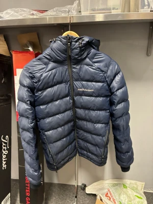 Blå dunjacka herr från Peak Performance  - Snygg blå dunjacka från Peak Performance med huva och dragkedja framtill. Jackan har två sidofickor med dragkedja och diskret logga på bröstet. Perfekt för kalla vinterdagar tack vare det tjocka dunfodret och den sportiga designen.