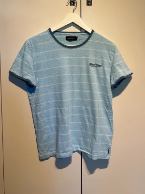 Ljusblå randig t-shirt East West - Snygg ljusblå t-shirt från East West med tunna vita ränder och broderad logga på bröstet. T-shirten har rund halsringning med mörkare blå kant och är kortärmad. Perfekt för en avslappnad och stilren look.