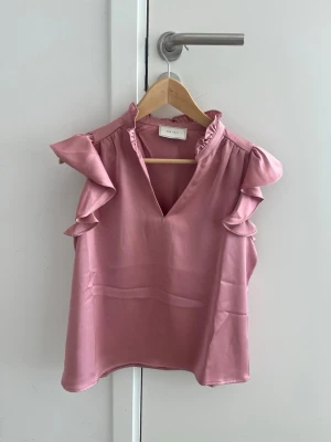 Neo Noir blus - Rosa blus från Neo Noir  ett glansigt, mjukt material. Blusen är ärmlös med volangdetaljer längs axlarna och har en liten ståkrage med v-ringning framtill. Använd endast en gång
