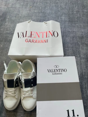 Valentino Garavani vita sneakers - Snygga vita sneakers från Valentino Garavani med bred svart rem över vristen och nitar på sidan. Skorna har klassisk låg siluett, vita snören och detaljer i läder. Perfekta för dig som vill ha en clean men ändå lyxig look.