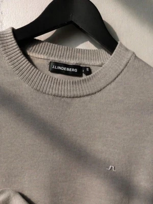 Beige tröja från J.Lindeberg - Snygg beige tröja från J.Lindeberg med rund halsringning och diskret logga broderad på bröstet. Tröjan har en klassisk passform och är perfekt för en stilren look. Materialet känns mjukt och bekvämt mot huden.