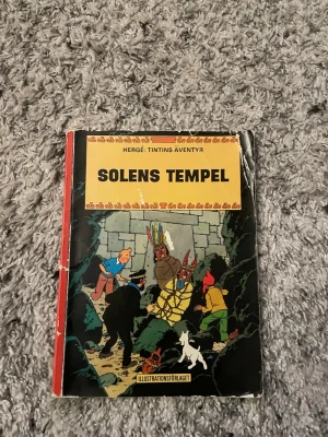 Tintins äventyr: Solens tempel - Säljer serieboken 'Solens tempel' från Tintins äventyr av Hergé. Boken har ett färgstarkt omslag med Tintin, Kapten Haddock och indianer, samt klassiska tecknade illustrationer. Omslaget är gult, rött och svart med detaljerade bilder och text.