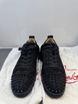 Christian Louboutin svarta spikes sneakers - Svarta sneakers i mocka från Christian Louboutin med svarta nitar över tån och klassisk röd sula. Skorna har svarta skosnören och låg profil. Perfekt för dig som vill sticka ut med en edgy och lyxig stil.